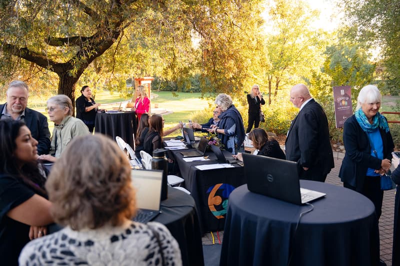 Guests mingle at tables set up on a sunny lawn under tall trees.
