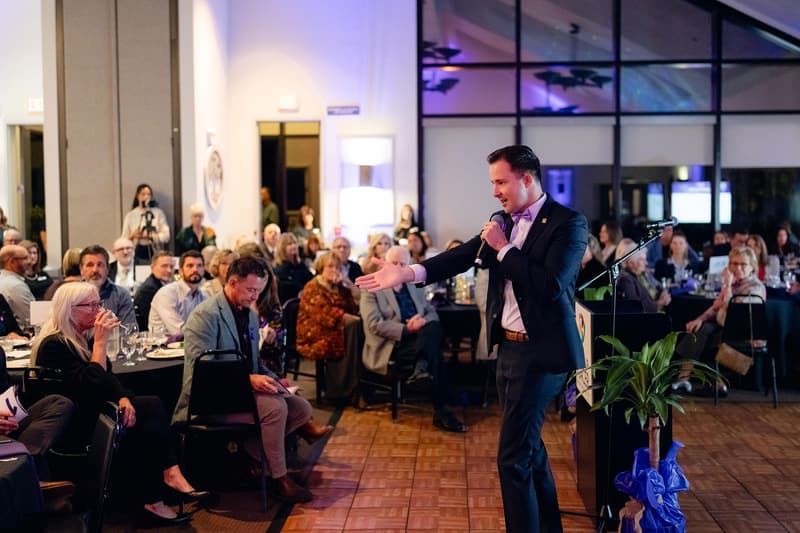 A host with a microphone speaks to guests seated around the room.