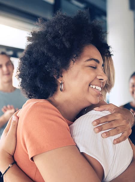 A smiling woman with curly hair hugs a friend.