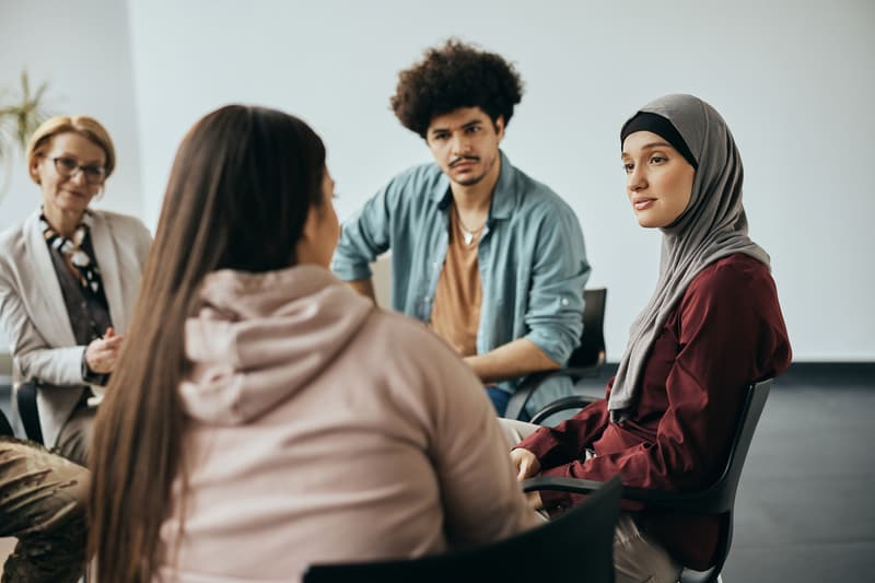 A small group sits in a circle of chairs during a counseling session; two women face the camera.