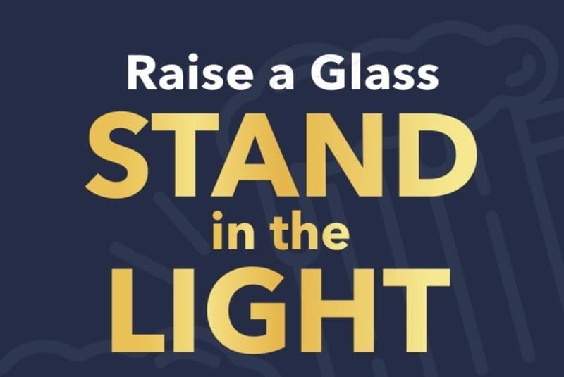 “Raise a Glass — Stand in the Light” in gold lettering on navy.